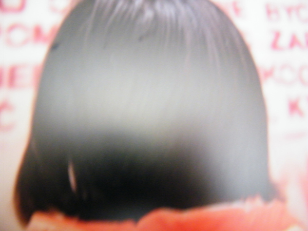 Kadr zdjęcia wypełnia sfotografowana od tyłu dziewczęca głowa. Na ciemne obcięte nad pomarańczowy kołnierz włosy poda jasna smuga światła. Na brzegach fotografii widoczne są rozmazane fragmenty napisów na murze, które odczytuje dziewczyna
