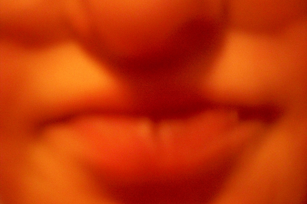 Zdjęcie przedstawia usta i fragment nosa autorki sfotografowane przez nią z bardzo bliska. Kontury są miękkie i płynne, a usta  rozmywają się w plamy różu i żółcieni